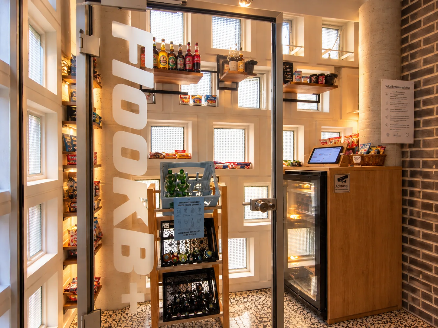 Blick durch die offene Glastür in den Selbstbedienungs-Kiosk mit Regalen voller Getränke und Snacks